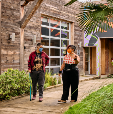 A person with a cane and a person with an abdominal binder walk along a path